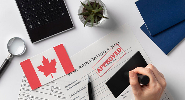 A person stamps a Canadian Visa Application form with APPROVED. In the background, a Canadian flag, calculator, magnifying glass, and two passports suggest a successful visa grant